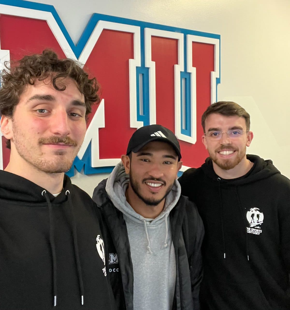 Trois jeunes hommes en sweat posent pour un selfie devant le grand logo rouge et bleu d'une université américaine