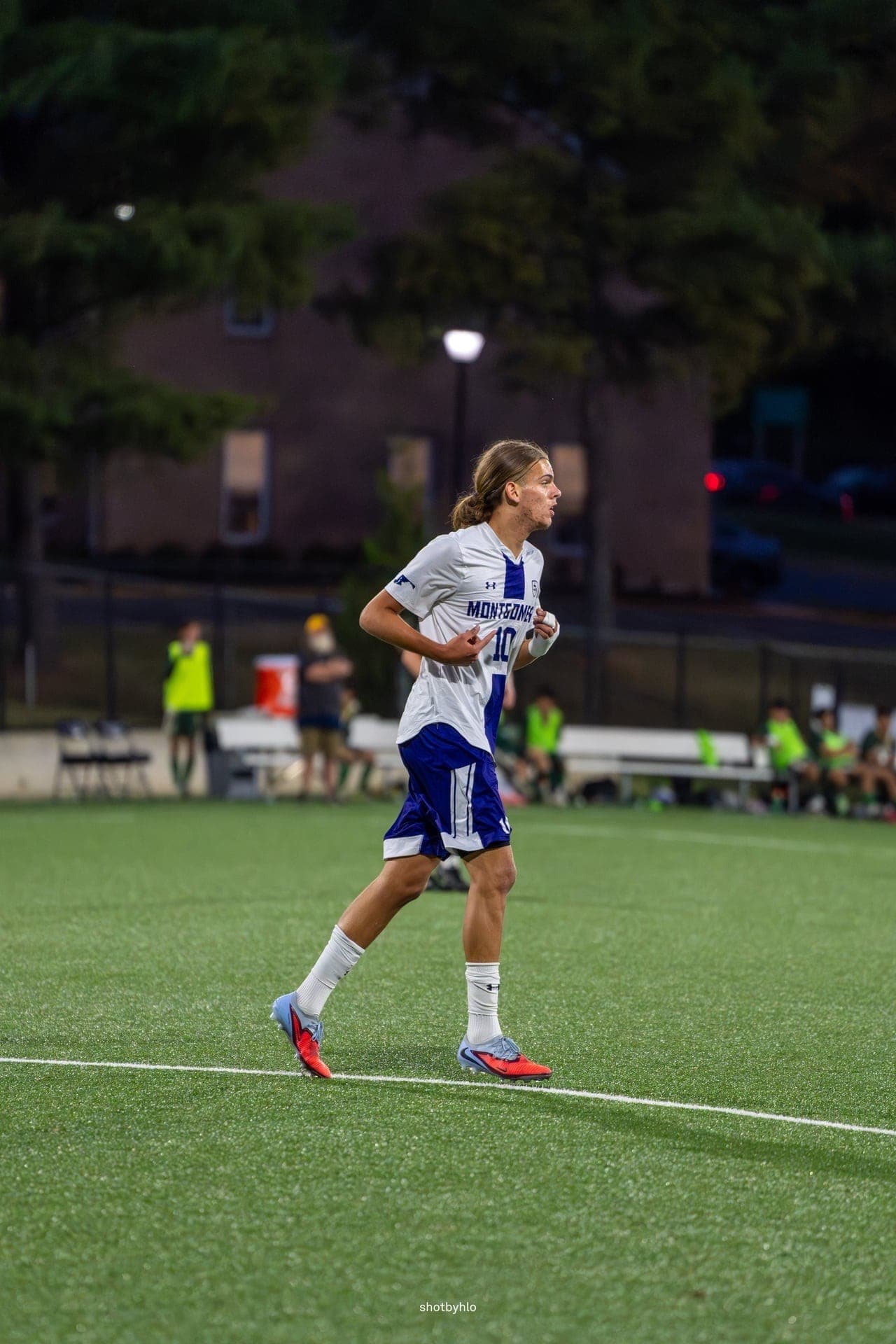 Un joueur en maillot blanc et bleu, numéro 10, court sur un terrain synthétique pendant un match de soccer en soirée