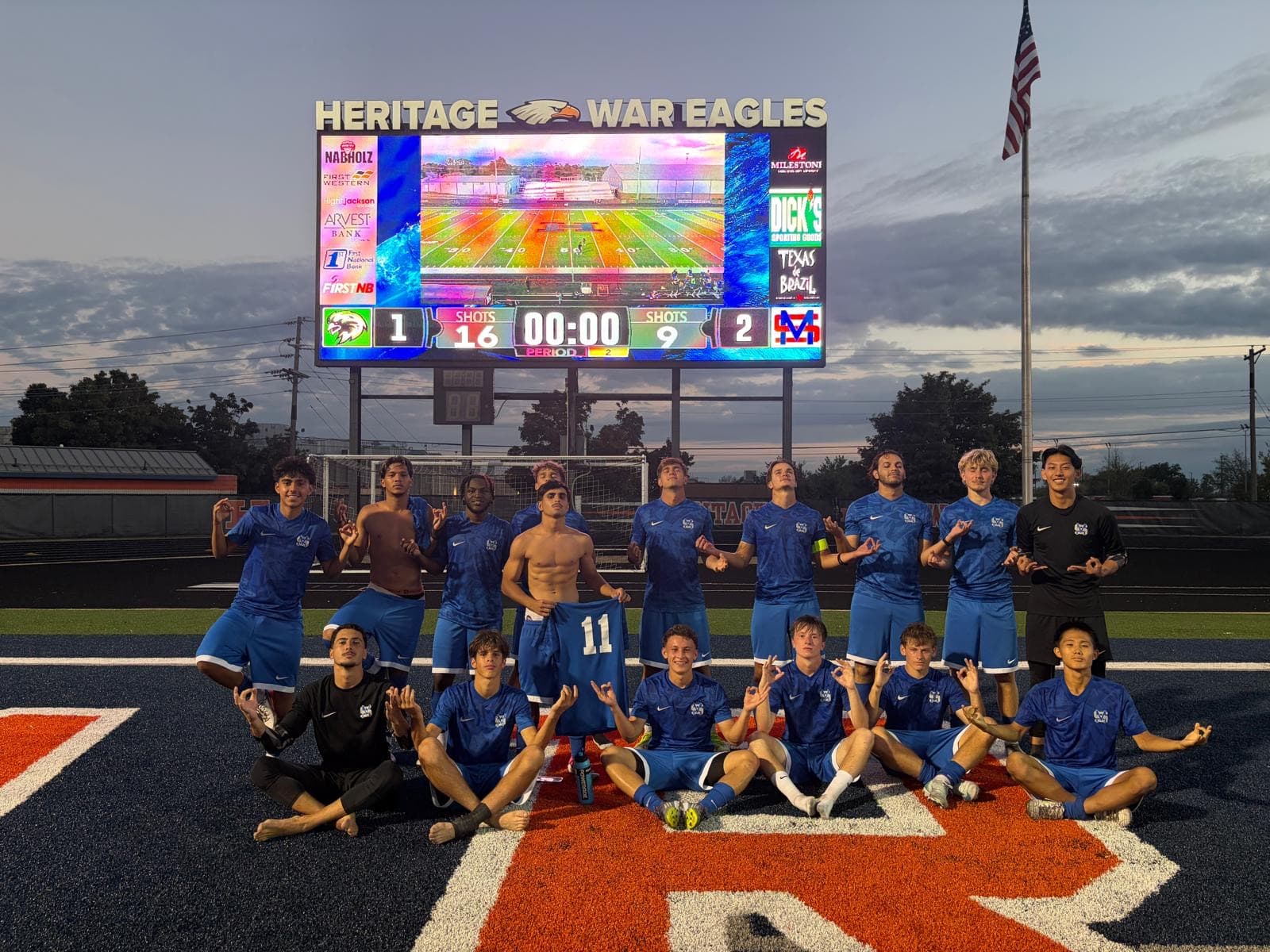 Une équipe universitaire de soccer en maillots bleus pose sur le terrain devant un tableau d'affichage, un soir de match aux États-Unis
